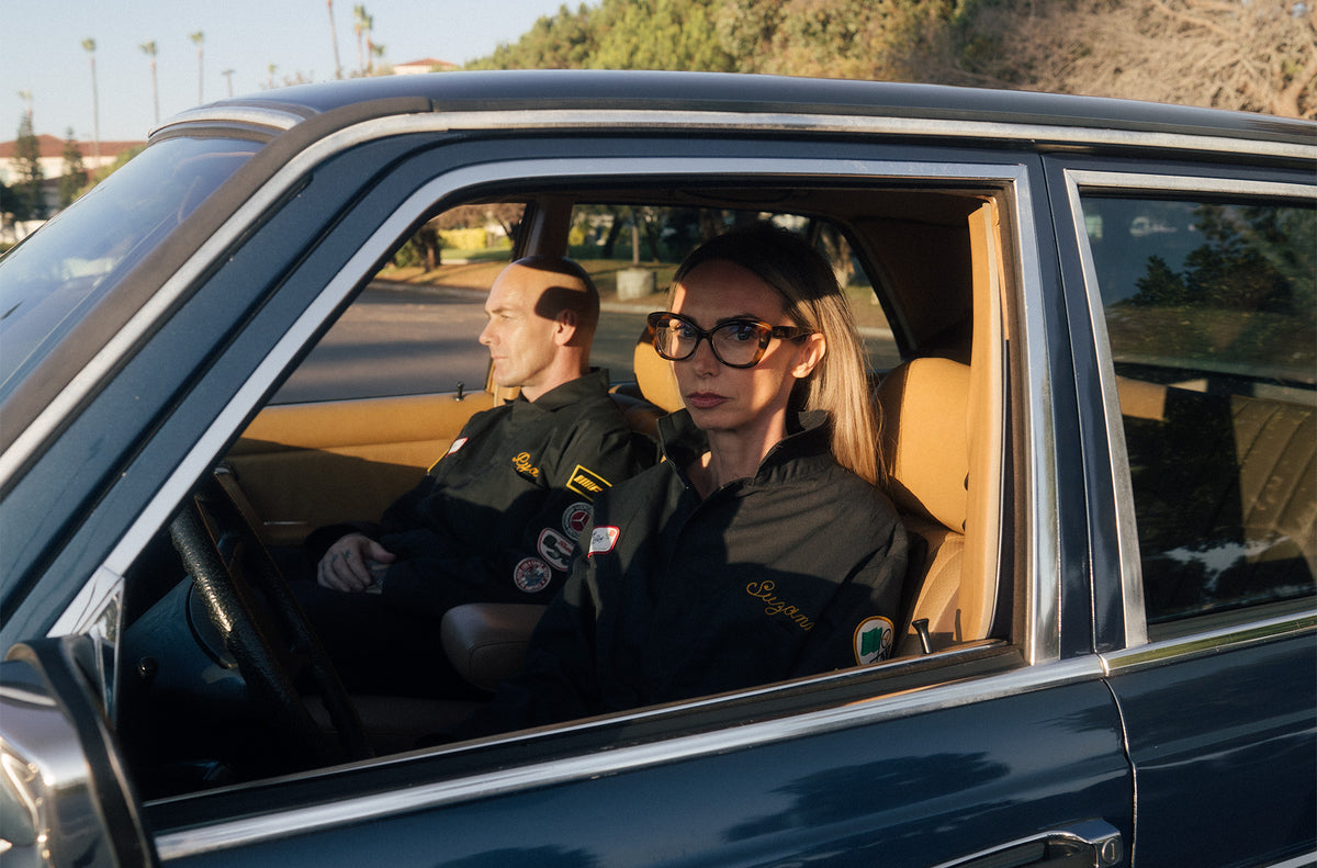 Two people in a car, one driving and the other sitting in the passenger seat, with a blurred outdoor background.