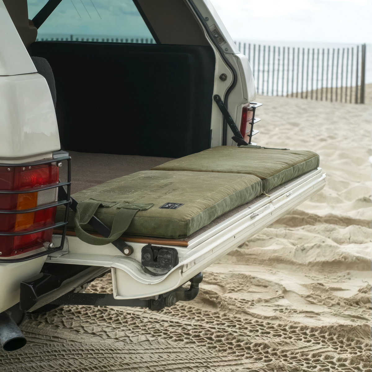 Two olive canvas tailgate cushions on the open tailgate of a white Range Rover Classic parked on the beach